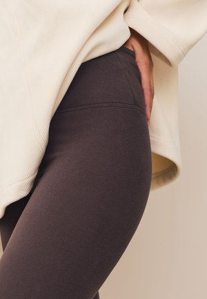 Prim-plan cu o persoană purtând colanți maro închis și un pulover crem supradimensionat, cu o mână sprijinită pe șold.