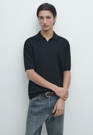 Jonge man met donker haar, gekleed in een zwart poloshirt met korte mouwen en een blauwe spijkerbroek, staand tegen een effen lichtgekleurde muur.