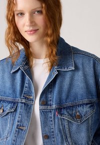 Modra jeans jakna z dvema prsna žepa, kovinskimi gumbi in klasičnim ovratnikom, nošena čez belo srajco z gladko teksturo.