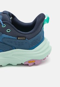 Tumma sininen vaelluskengässä, jossa on verkkokangas- ja synteettinen päällinen, mintunvihreä välipohja, violetti ulkopohja ja Gore-Tex- sekä Vibram-brändäys.