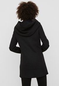 Cappotto nero con cappuccio drappeggiato, vestibilità su misura e tessuto liscio, con cuciture verticali per il supporto e un taglio di lunghezza media.