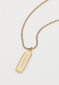 Gyldent metal halskæde med en rektangulær vedhæng indgraveret med "LACOSTE," der har en glat overflade og en delikat kæde.