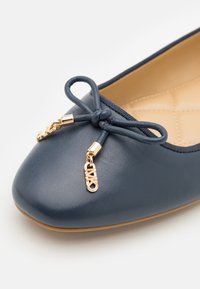 Ballerine in pelle blu scuro con punta arrotondata, caratterizzata da un fiocco decorativo e dettagli in hardware dorato. L'interno presenta un motivo trapuntato.