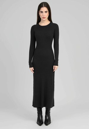 Femme portant une robe midi en maille côtelée noire à manches longues et des bottes noires à bout pointu, debout devant un fond gris clair.
