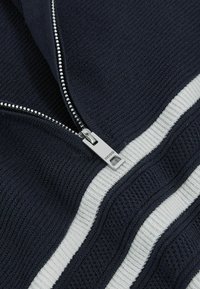 Mornarska zip-up dukserica s teksturiranim pletenjem, dva bijela horizontalna pruga i srebrni patent zatvarač s detaljem logotipa.