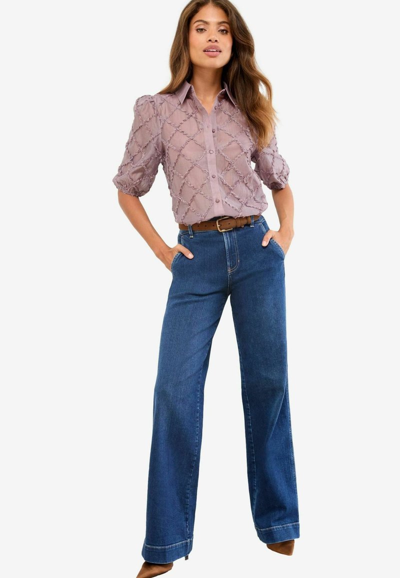 Lyse lilla bluse med tekstureret mønster og pufærmer, parret med højhalsede blå vide jeans og et brunt bælte.