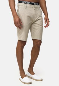 Shorts beige con un patrón texturizado, bolsillos frontales y una cinturilla negra con marca. Combinados con zapatillas casuales blancas.
