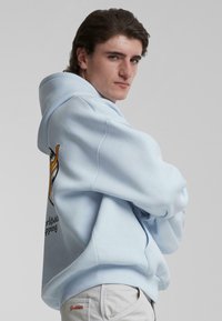 Felpa leggera blu in misto cotone, con una stampa grafica sulla schiena e polsini a coste. Abbinata a pantaloni beige.
