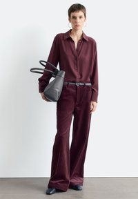 Maroon button-up shirt, wide burgundy corduroy pants, black belt, and a soft grey handbag with dual handles, set against a neutral background.