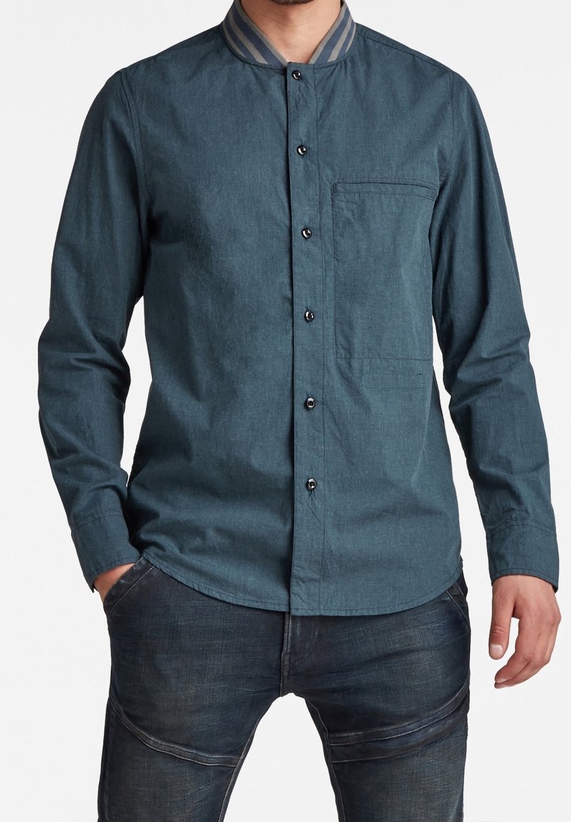 Camisa de botón en algodón color teal oscuro con un collar a rayas grises, un bolsillo en el pecho y botones negros; presenta mangas largas y un dobladillo curvado.