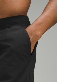 Svarta shorts med en slät textur, med sidofickor och resårmidja. Närbild på fickområdet med hudton synlig.