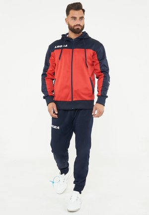 Uomo che indossa una giacca con cappuccio rossa e blu navy con zip e pantaloni della tuta blu navy con scarpe da ginnastica bianche, in piedi su uno sfondo bianco.
