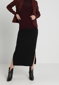 Blazer à carreaux bordeaux sur un top en maille bordeaux, associé à une jupe midi noire avec fentes latérales, et des bottines noires avec fermetures éclair.
