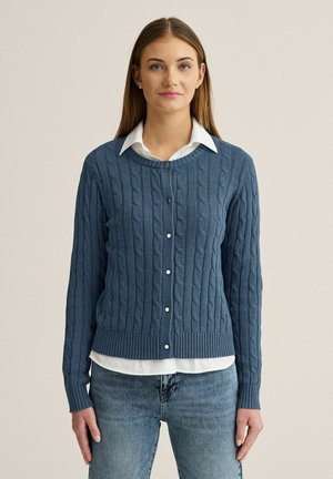 Kvinde stående, iført en blå kabelstrikket cardigan over en hvid skjorte med krave og blå jeans, vendt fremad mod en ensfarvet baggrund.