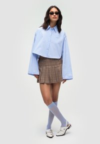 Camicia azzurra chiara tagliata, con maniche oversize, abbinata a una gonna plissettata a quadri marrone, calze alte fino al ginocchio azzurre e sandali bianchi.