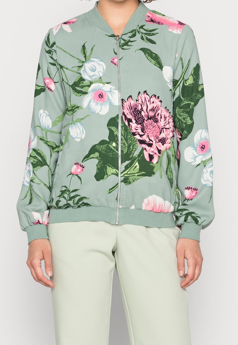 Veste à fermeture éclair vert clair avec un grand motif floral rose et blanc, portée avec un pantalon beige clair.