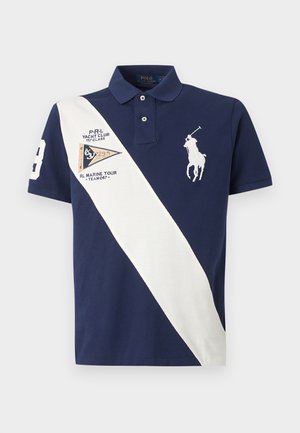 Νέος άνδρας με ναυτικό μπλε πόλο με λευκή διαγώνια ρίγα, μπεζ παντελόνι cargo και λευκά αθλητικά, όρθιος με τα χέρια στις τσέπες.