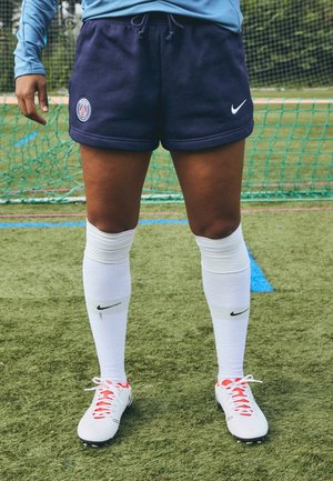 Benen die marineblauwe Paris Saint-Germain shorts dragen, witte Nike kniehoge sokken en witte voetbalschoenen, staand op een grasvoetbalveld.