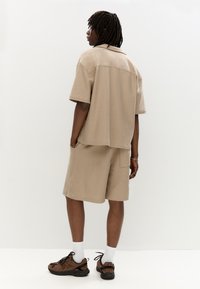 Chemise beige à manches courtes et short en tissu texturé, avec une coupe décontractée. Portée avec des baskets marron et des chaussettes blanches.