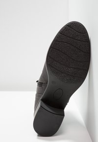 Semelle de chaussure noire avec des motifs d'accroche texturés et le numéro de taille 37 visible, placée debout contre un fond blanc en coin.