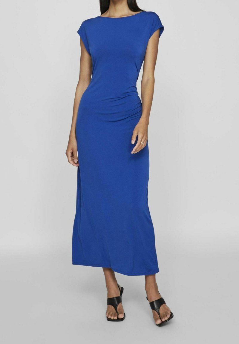 Robe bleue arrivant au genou en tissu lisse, avec un col rond, des manches courtes et des fronces sur les côtés. Portée avec des sandales noires.