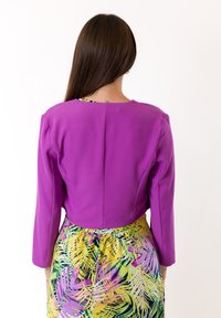 Giacca corta viola con maniche lunghe e design strutturato. Abbinata a una gonna decorata con motivi gialli e verdi che presentano stampe di foglie di palma.