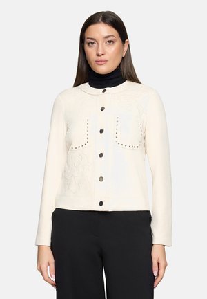 Ivoorwitte jas met lange mouwen, voorzien van twee borstzakken, decoratieve metalen studs en een bloemenreliëf langs de linkerkant.