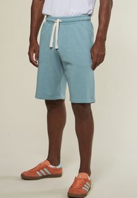 Mann in hellblauen Joggingshorts mit Kordelzug und orangefarbenen Sneakern mit hellblauen Streifen, vor einem neutralen Hintergrund stehend.