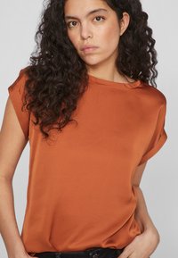 Top corto arancione ruggine in tessuto morbido con vestibilità rilassata e scollatura tonda. Design minimalista con dettagli delle maniche arrotolate.