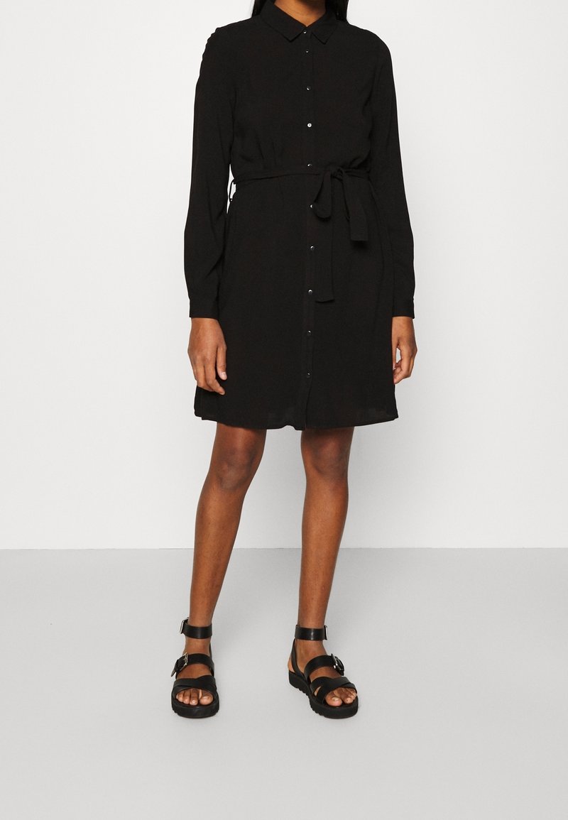 Femme portant une robe noire à manches longues boutonnée avec ceinture à la taille et des sandales noires à boucle, debout devant un fond blanc uni.