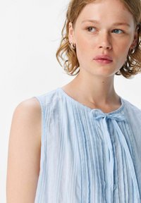 Una joven con el cabello corto y rizado lleva una blusa sin mangas de color azul claro y con pliegues, mirando pensativa hacia un lado contra un fondo sencillo.