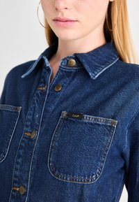 Camisa de mezclilla azul oscuro con cuello, que presenta dos bolsillos frontales y botones de latón. Incluye costuras visibles y una etiqueta con el logo.