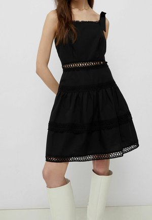 Robe noire à volants avec un décolleté carré, détail de nœud sur une épaule, garniture brodée et ourlet texturé. Le mannequin porte des bottes blanches jusqu'aux genoux.