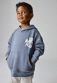 Ragazzino in felpa blu oversize con le mani nelle tasche che guarda di lato su uno sfondo grigio semplice.