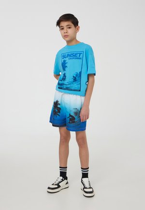 T-shirt in cotone blu con grafica "SUNSET CALIFORNIA" e palme. Pantaloni da bagno blu sfumati abbinati con scena di surf. Sneakers in bianco e nero.