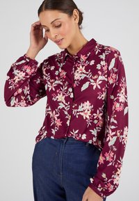 Camisa de botones de color burdeos con estampado floral, hecha de una tela suave, que presenta patrones de flores en tonos rosa y verde, y con mangas largas con puños abotonados.
