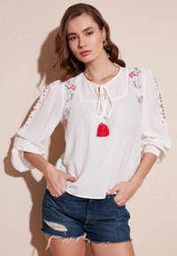 White blouse with floral embroidery in red and green, featuring a tie neck and a red tassel. Paired with distressed denim shorts.