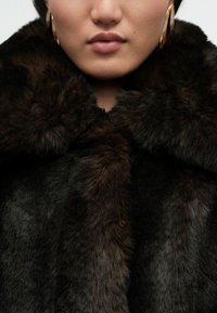 Ψεύτικο παλτό από γούνινο υλικό σε πλούσιο σκούρο καφέ χρώμα με αφράτη υφή. Διαθέτει φαρδύ γιακά και κομψό, στενό σχεδιασμό.