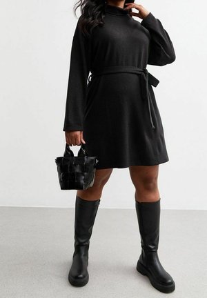 Femme en robe noire ceinturée à manches longues, tenant un sac à main noir tressé, portant des bottes noires montantes, debout sur un sol gris.