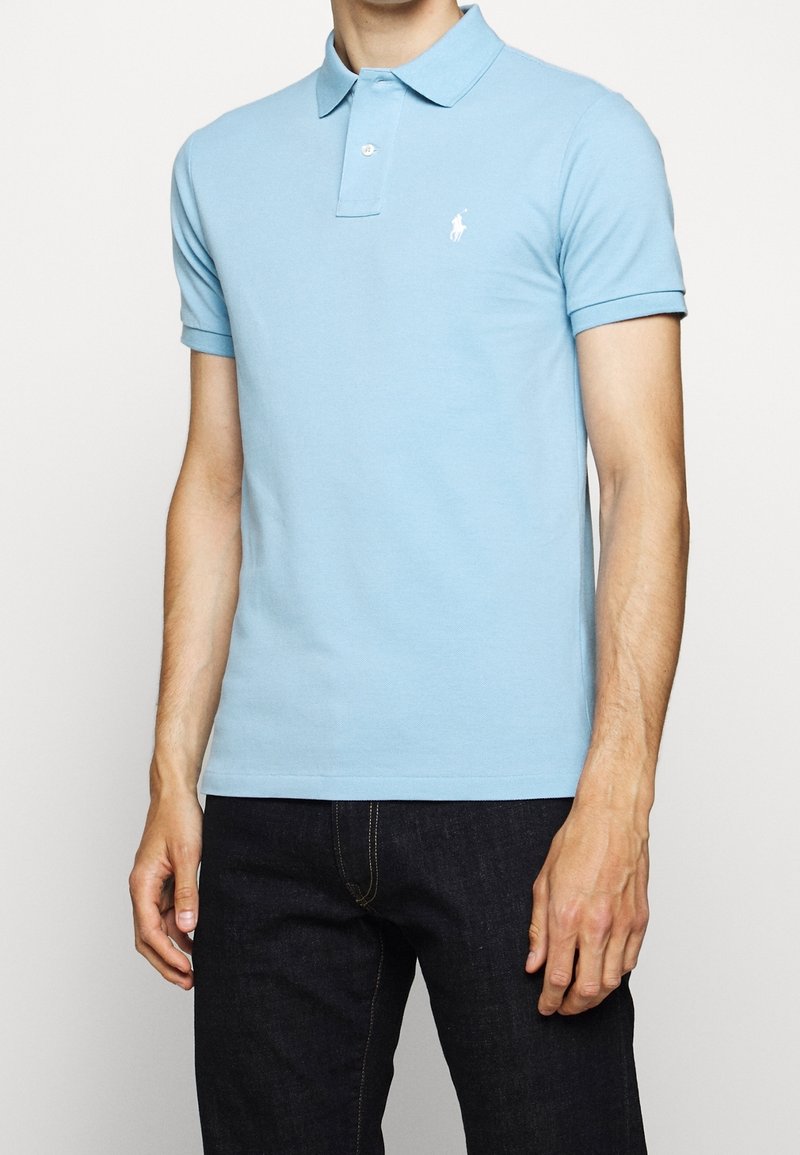 Man som bär en ljusblå kortärmad pikétröja med en liten vit logo på bröstet, i kombination med mörkblå jeans, står mot en enkel bakgrund.