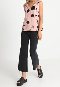 Nainen, jolla on yllään hihaton vaaleanpunainen kukkakuvioinen paita, mustat lyhytlahkeiset liehuhousut ja mustat korolliset liukusandalit kävelemässä.