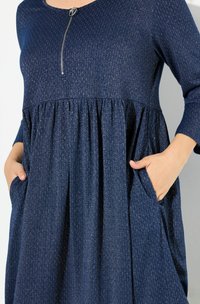 Tummansininen mekko tekstuuripinnalla, jossa on pyöreä pääntie, vetoketjuyksityiskohta ja kerätty vyötärö. Sisältää sivutaskut ja pitkät hihat.