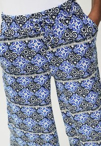 Pantalons légers à motifs bleu et blanc avec un design floral, ceinture élastique et poches latérales. Présente des accents noirs contrastants.