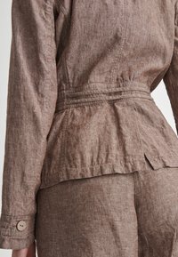 Hnědá lněná bunda s peplum designem, s jemnou texturou, nařaseným pasem a zapínacími manžetami.