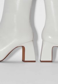 Botas de cuero blanco hasta la rodilla con un tacón cuadrado, que presentan una textura suave y acentos marrones contrastantes en la base del tacón.
