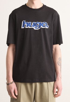 Osoba nosząca czarną koszulkę z "hugo." w niebieskim wzorzystym tekście oraz jasnobeżowe spodnie, z tatuażem na lewym przedramieniu.
