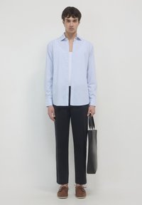 Hellblau gestreiftes Hemd mit Knöpfen, schwarze Hose, braune Schnürschuhe, hält eine schwarze Tragetasche. Schlichtes Design, lässige Passform.