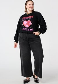 Zwart sweatshirt met lange mouwen en roze hartmotief, gecombineerd met losse zwarte spijkerbroek en zwarte muiltjes met hak. Accenten van zilveren sieraden.