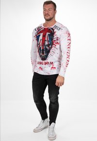 Witte longsleeve t-shirt met een grafisch ontwerp van een schedel en de Union Jack, met rode spattenprint. Draagt het met zwarte gescheurde jeans en witte sneakers.