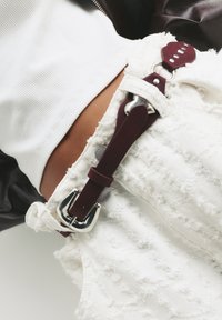 Fehér, megviselt farmer sort, rojtos szegéllyel, burgundi bőrszíjjal és ezüst hardver díszítéssel. Texturált és laza kialakítás.